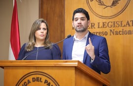 Una mujer de mediana edad vestida con una camisa azul de mangas abullonadas. A su lado un hombre joven con barba, camisa blanca, saco azul, que levanta un dedo índice; ambos detrás de un atril con el escudo de Paraguay. A la izquierda una bandera paraguaya. De fondo un escudo del Poder Legislativo en la pared revestida con madera.