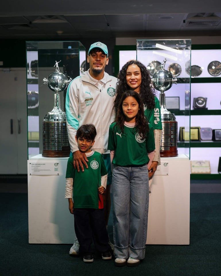 Hombre con chaqueta blanca y gorro verde sonríe junto a familia en ambiente colorido con trofeos del Palmeiras.