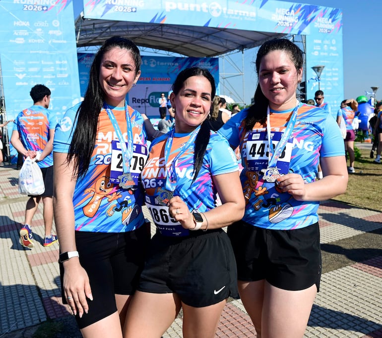 Tres mujeres sonrientes en camisetas azules con detalles naranjas, sosteniendo medallas, en un evento deportivo en el exterior.