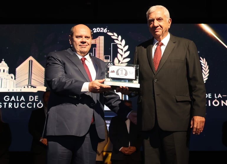 Hombre de traje azul oscuro y corbata roja sostiene un trofeo, mientras otro con cabello canoso viste un traje negro con corbata a rayas rojas.