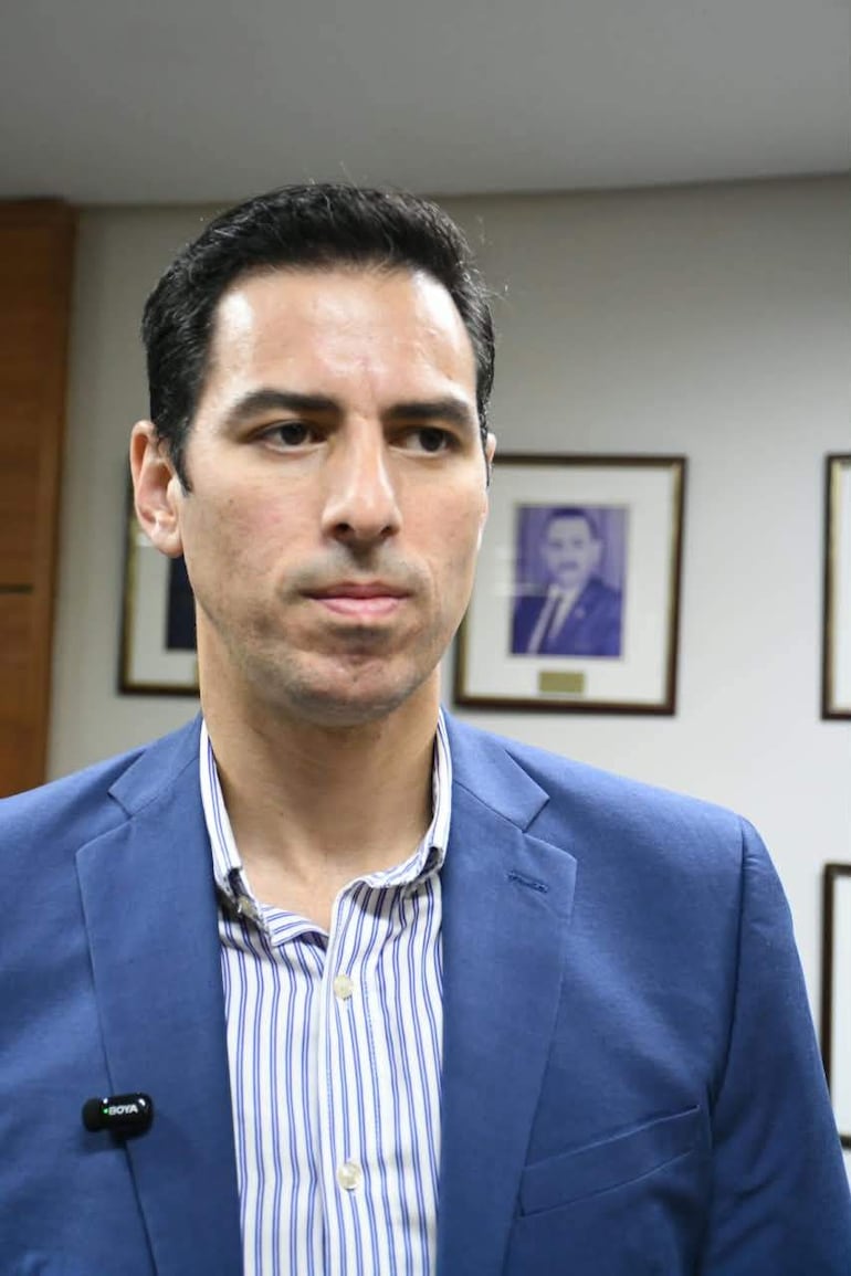 Alvaro Grau de pie en oficina, vistiendo blazer azul y camisa de rayas, con expresiones tranquilas y fondo de retratos enmarcados.