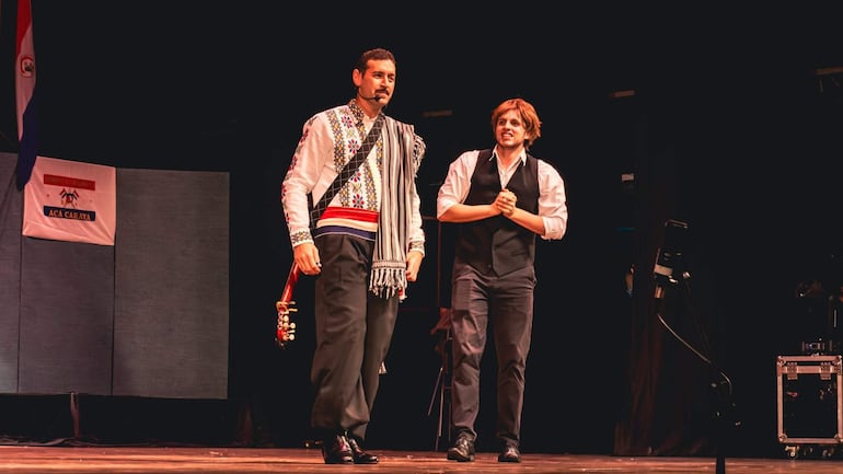 Luis Alberto del Paraná en camisa tradicional y poncho con guitarra, mientras un hombre en chaleco negro lo observa atentamente.