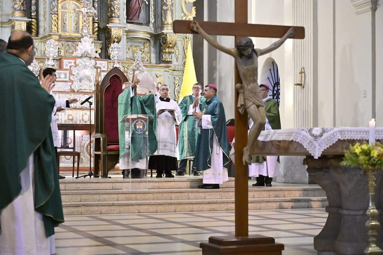 Sacerdote en vestimenta verde sostiene un documento, mientras obispo con sombrero púrpura lo observa en una ceremonia religiosa solemne.