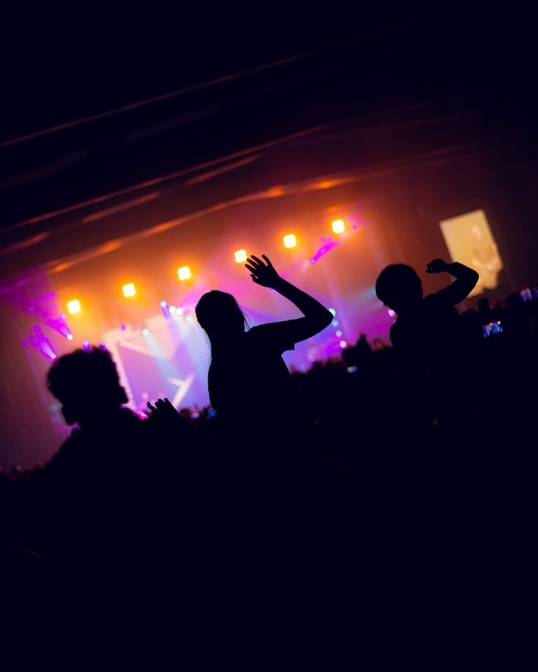 Asistentes al concierto con manos levantadas, ante luces moradas y naranjas en un ambiente vibrante.