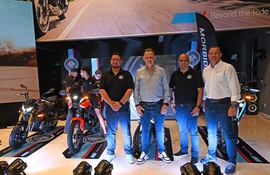 Cuatro hombres vestidos de negro presentan motocicletas en exhibición, con banner de Morbidelli al fondo y luces encendidas.