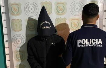 Hombre encapuchado y policía de espaldas en un entorno interior con insignias policiales visibles.