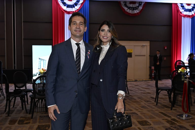 Hombre en traje oscuro con medalla y mujer en conjunto oscuro sonriendo en decoración azul, blanco y rojo.