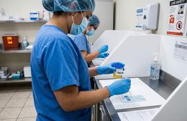 Dos profesionales de la salud en uniformes azules y guantes, uno sostiene un frasco amarillo mientras el otro utiliza un equipo de laboratorio.