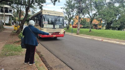 Mujer de espaldas con abrigo azul y pantalones negros con franjas rojas, extendiendo su brazo hacia un autobús.