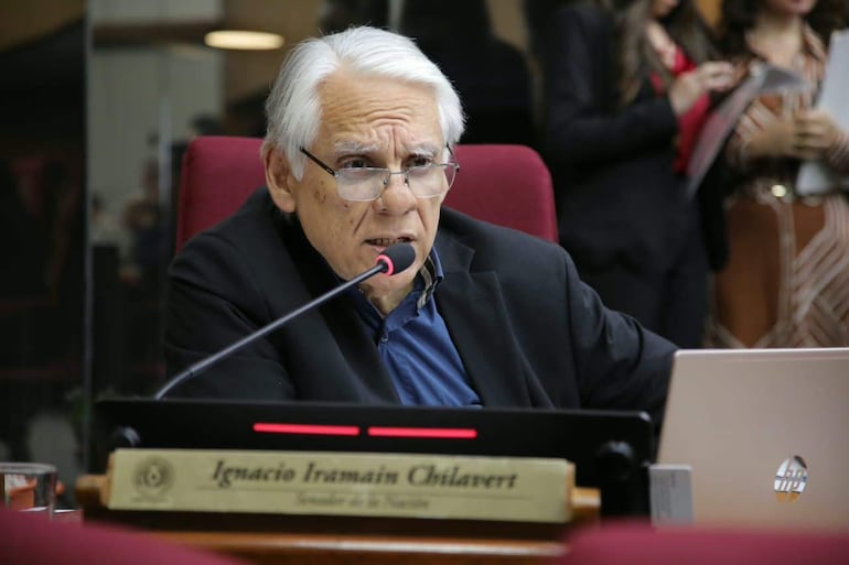 Ignacio Iramain Chilavert con cabello blanco y gafas, sentado en casaca negra, habla junto a un micrófono y papeles.