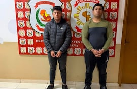 Dos hombres detenidos, uno en abrigo negro y otro en camisa negra, de pie bajo custodia en una estación de policía, rodeados de armas.