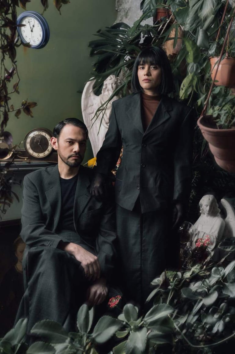 Hombre con traje oscuro y mujer de pie en conjunto similar, rodeados de plantas y decoración en un ambiente acogedor.