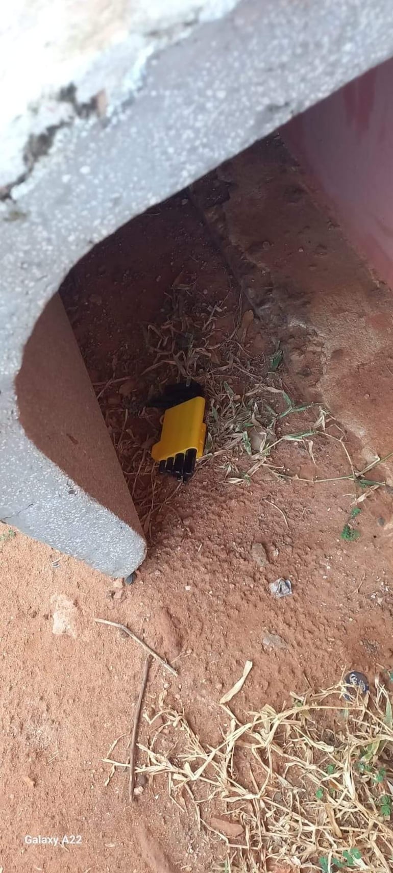 Un cargador amarillento parcialmente cubierto por un bloque de cemento en suelo de tierra y hierba seca.