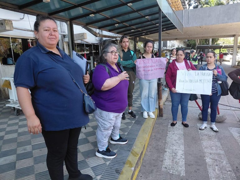 Seis personas, principalmente mujeres, sostienen carteles en protesta, una mujer en morado habla y otra escucha atentamente, ambiente urbano.