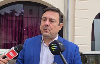 Enrique Buzarquis con sacón azul, siendo entrevistado por reporteros con micrófonos en un ambiente informal.