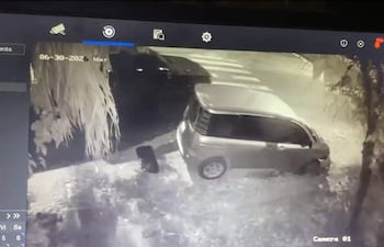 Coche oscuro avanzando en una calle con un vehículo plateado estacionado, en un entorno nocturno sin personas presentes.