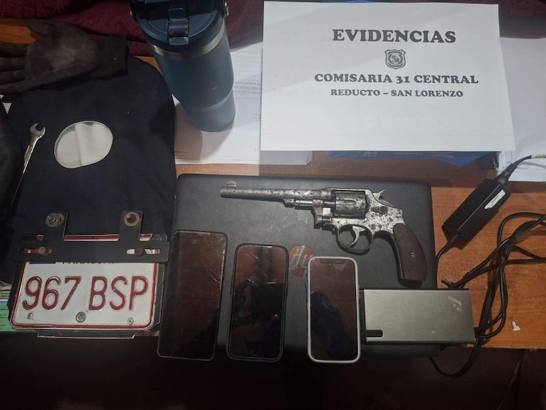 Revólver sobre mesa de madera, junto a teléfonos móviles, guante negro y documentación de evidencia en un ambiente cerrado.