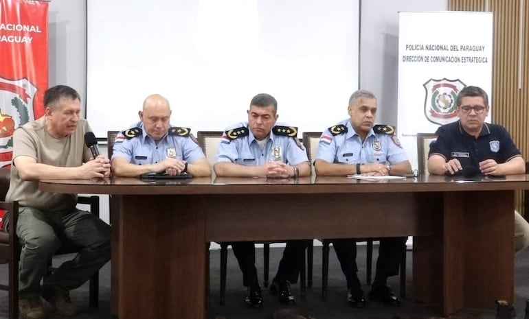 Cinco hombres en conferencia de prensa, visten camisas claras con insignias y micrófono, en un ambiente formal con logos de policía.
