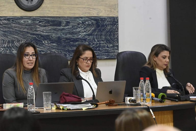 Tres mujeres en mesa de trabajo: una habla con blusa gris, otra concentra en su laptop con blusa blanca y chaqueta negra, y la tercera revisa documentos.