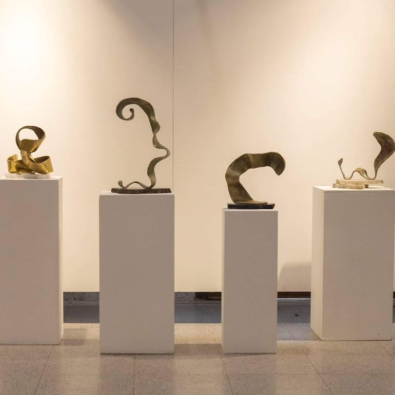 Cuatro esculturas abstractas en pedestales blancos, con acabados dorados y tonos neutros, en una galería desierta.