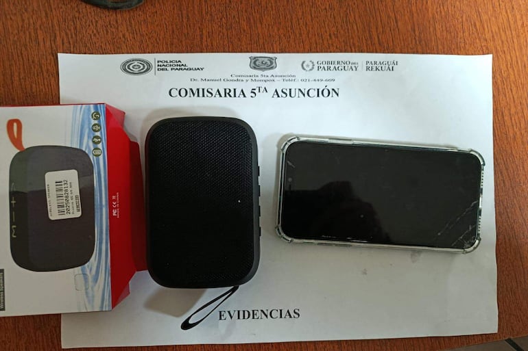 Mesa con un altavoz portátil negro y un teléfono móvil dañado sobre documentos policiales en comisaría.