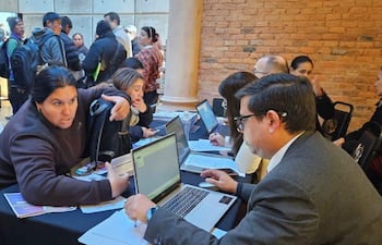 Mujer de cabello oscuro gesticula mientras habla con un hombre concentrado en su laptop en un espacio de atención al público.