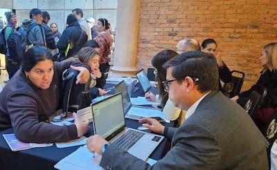 Mujer de cabello oscuro gesticula mientras habla con un hombre concentrado en su laptop en un espacio de atención al público.