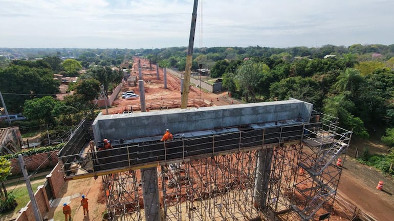 Cuatro trabajadores con cascos en obra de construcción, uno sentado sobre una estructura, rodeados de materiales y postes de concreto.