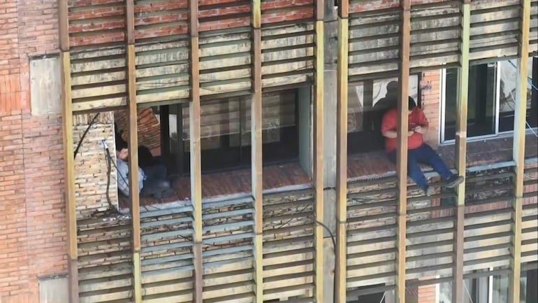 Hombre de camiseta roja sentado en la ventana de un edificio en construcción, con un teléfono móvil en la mano.