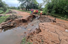 Cinco personas en un camino de tierra lleno de barro, rodeados de vegetación y un puente de madera en mal estado.