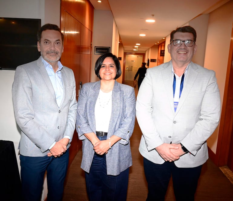 Tres asistentes en chaquetas formales sonríen en un evento, posando en un ambiente interior elegante.