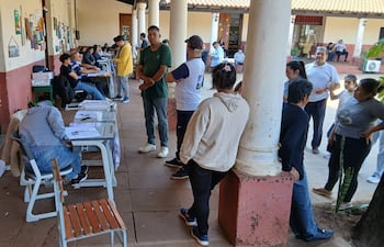 Hombres interactúan en centro de votación externo, algunos con documentos y visibles preocupaciones mientras completan formularios.