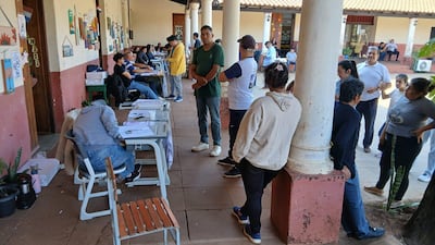 Hombres interactúan en centro de votación externo, algunos con documentos y visibles preocupaciones mientras completan formularios.