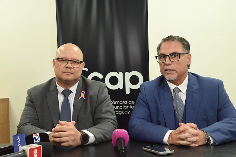 Oscar Lovera con chaqueta gris y gafas, sentado junto a un hombre de traje azul claro en una conferencia.