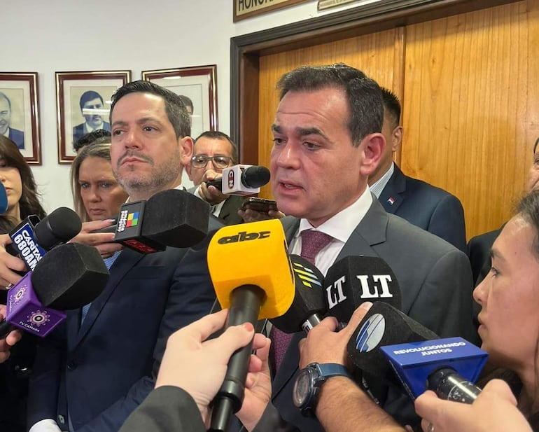 Rubén Ramírez en traje gris hablando a periodistas. Raúl Latorre lo acompaña, rodeados de varios reporteros en un ambiente formal.