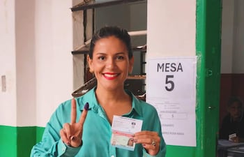 Noelia Díaz sonríe, viste blusa verde y pantalones blancos, sostiene su cédula y hace un signo de paz en un centro de votación.