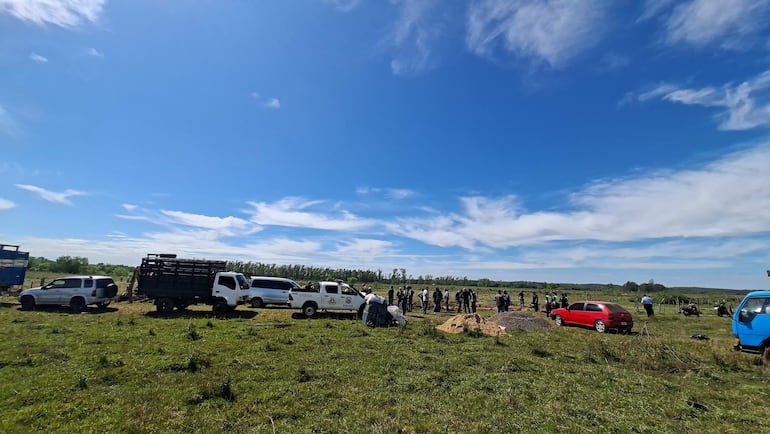 Un grupo de 20-30 personas discuten en terreno con pasto, algunos visten camisetas oscuras y vehículos varios al fondo.