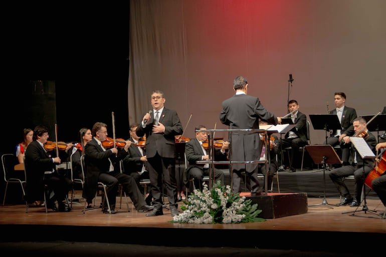 Músicos en trajes oscuros, un hombre con micrófono dirigiendo, y una mujer en vestido colorido observa atentamente durante el homenaje.