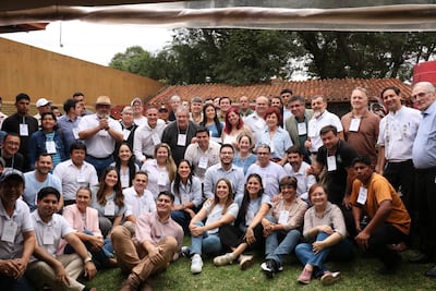 Miembros del partido Patria Querida sonríen y conversan en un ambiente al aire libre, con identificaciones visibles. Fondo amarillo y techado con tejas.
