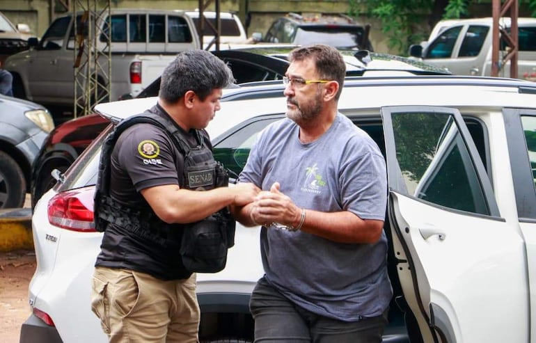 Hombre con camiseta gris y cadenas en las muñecas es sujetado por un agente de seguridad con chaleco de la Senad cerca de un coche blanco.