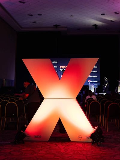Hombre en camiseta oscura ajusta equipo frente a un gran símbolo iluminado de TEDx en un auditorio.