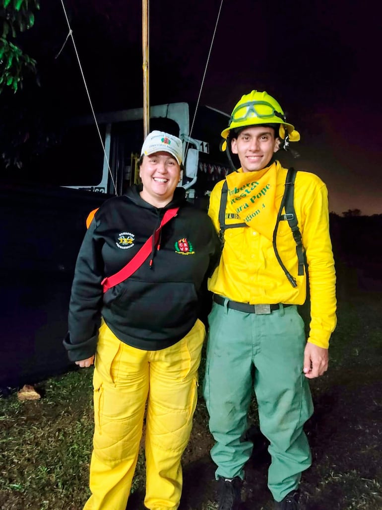 Lorena Canan en chaqueta negra y gorra sonríe junto a hombre en uniforme amarillo, en un ambiente exterior con poca iluminación.