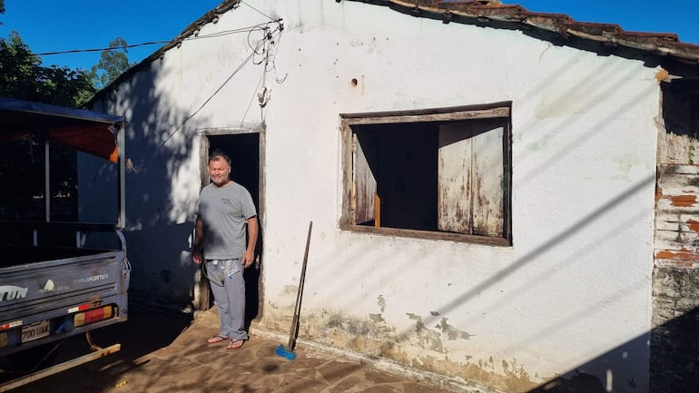 César Villalba posando de pie en la entrada de su vivienda, casa de paredes blancas y techo de tejas, lugar que iba a rifar para costear sus tratamientos médicos y cuidados de su familia en San Juan Bautista.