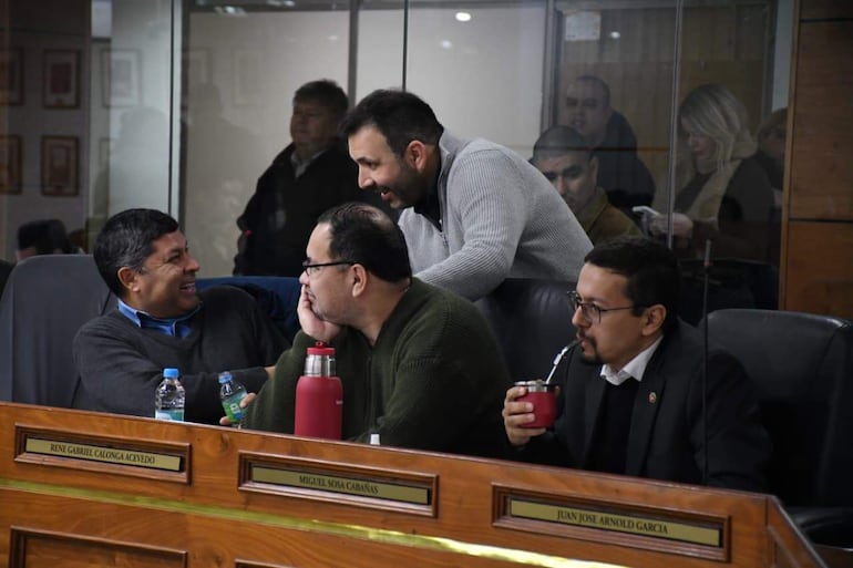 Cesar Escobar Ceres de pie gesticulando, mientras Miguel Sosa ríe a la izquierda, en ambiente de reunión con mesas de madera.
