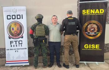 Hombre de camiseta clara en custodia de dos agentes de la SENAD, operativo con pancartas de seguridad en fondo.