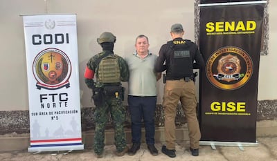 Hombre de camiseta clara en custodia de dos agentes de la SENAD, operativo con pancartas de seguridad en fondo.