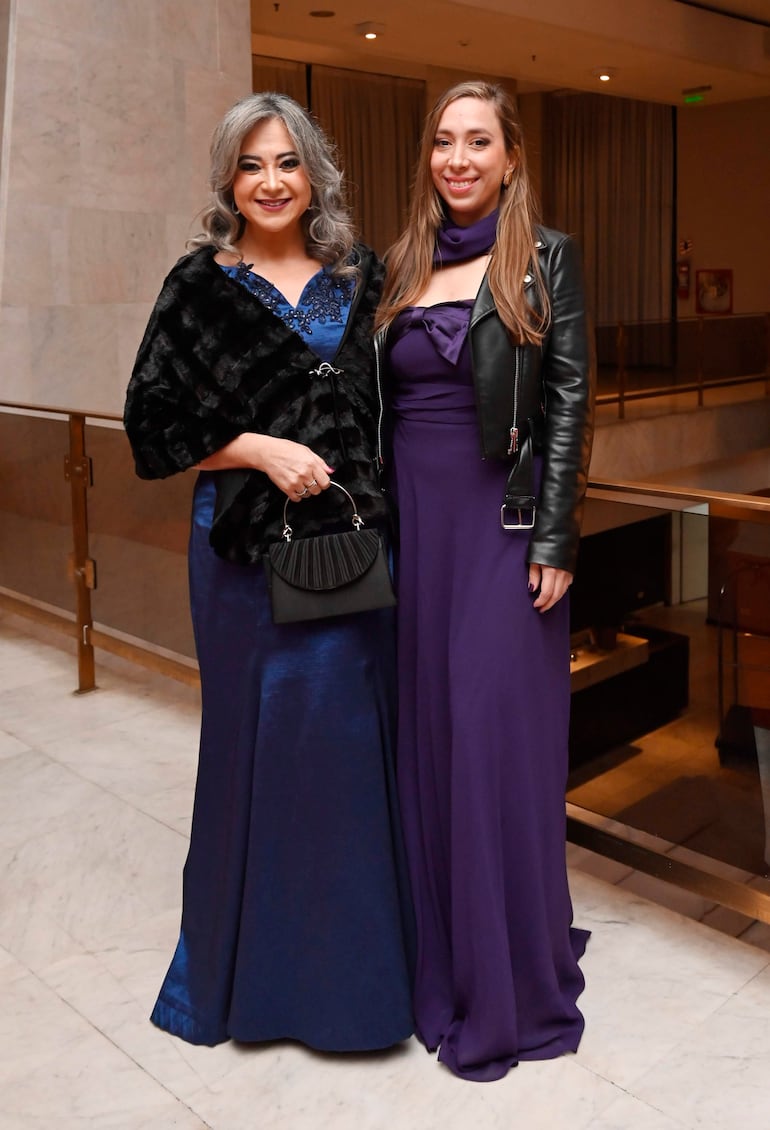 Dos mujeres sonrientes, una con vestido azul oscuro y abrigo negro, otra con vestido morado y chaqueta de cuero, en un entorno festivo.