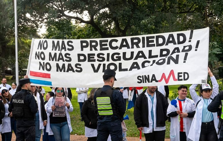 Médicos en batas blancas sosteniendo un banner con demandas, rodeados de policías en un ambiente de protesta.