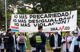 Médicos en batas blancas sosteniendo un banner con demandas, rodeados de policías en un ambiente de protesta.