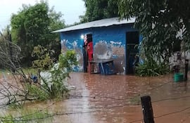 Escena de inundación en Carapeguá, con edificio azul afectado y objetos como sillas y un balde anegados.
